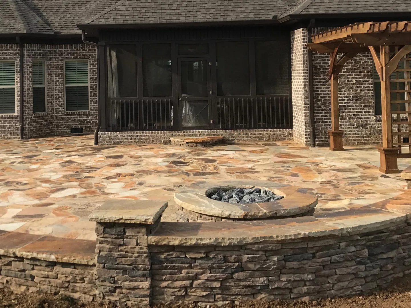 A round stone fire pit set in a curved stacked stone seat wall on a flagstone patio behind a brick house, outdoor fireplaces and fire pits