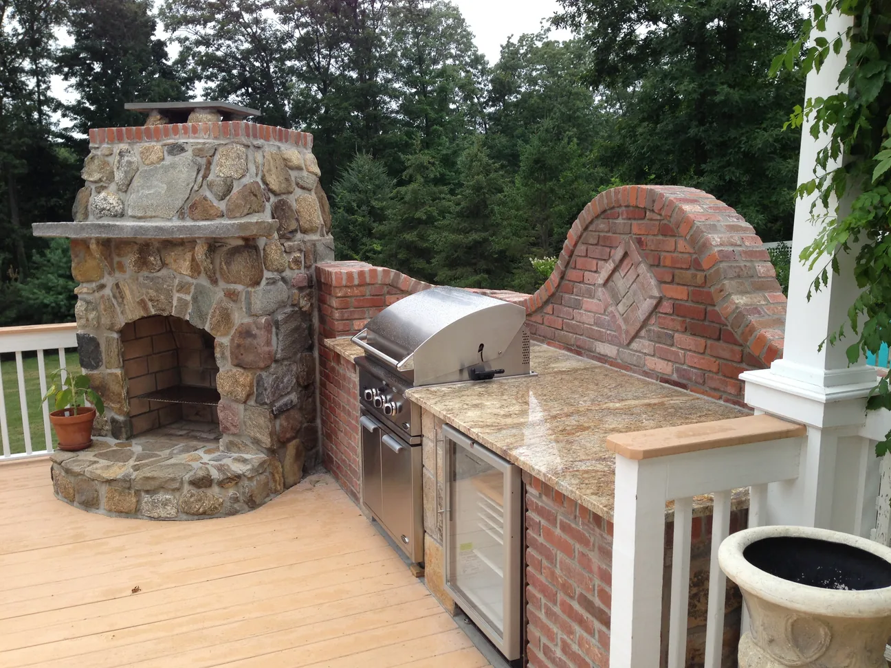 A brick grill island with a granite counter beside a fieldstone wood oven on a deck, outdoor kitchens