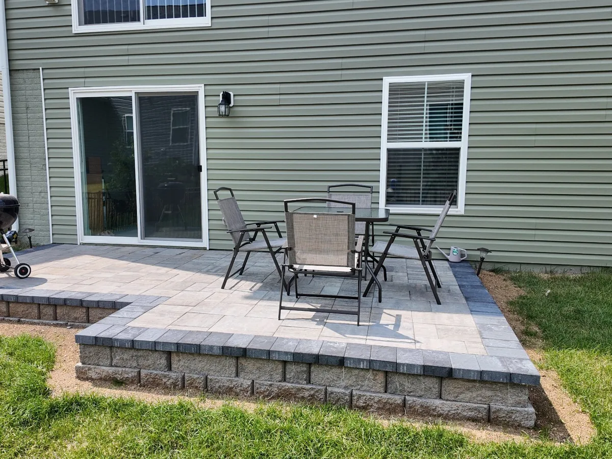 A raised paver patio on a block base with a grill and patio chairs behind a house, outdoor kitchens