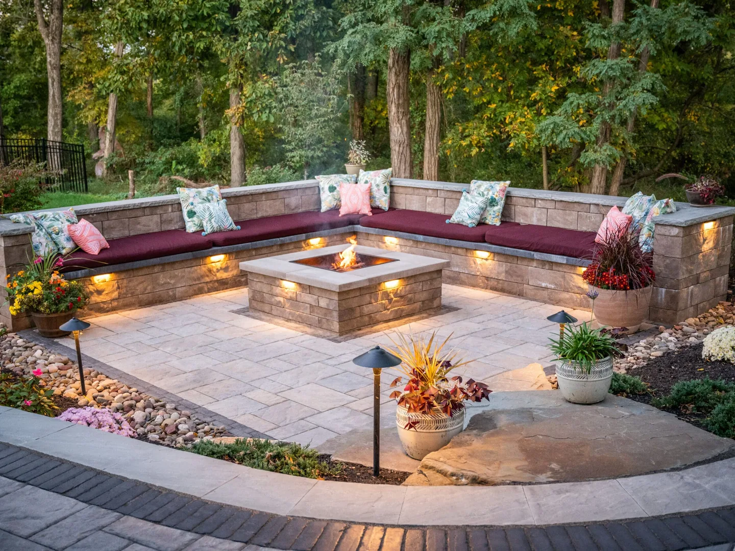 A sunken paver patio with a U shaped block seat wall around a block fire pit at dusk, outdoor kitchens