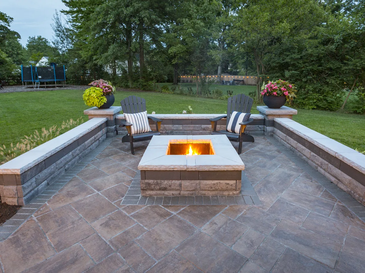 Low block seat walls with stone caps around a square fire pit on a paver patio, outdoor kitchens