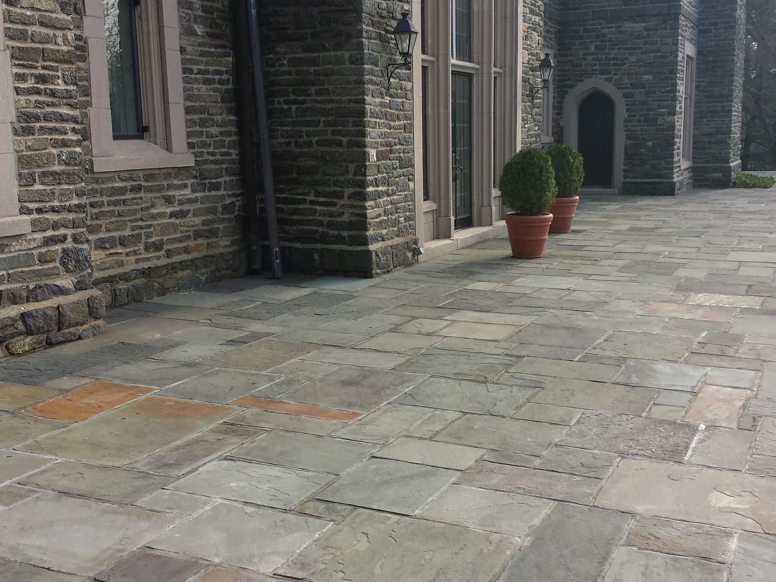 A bluestone patio laid along the stone walls of a house with potted boxwoods, brick and stone patios