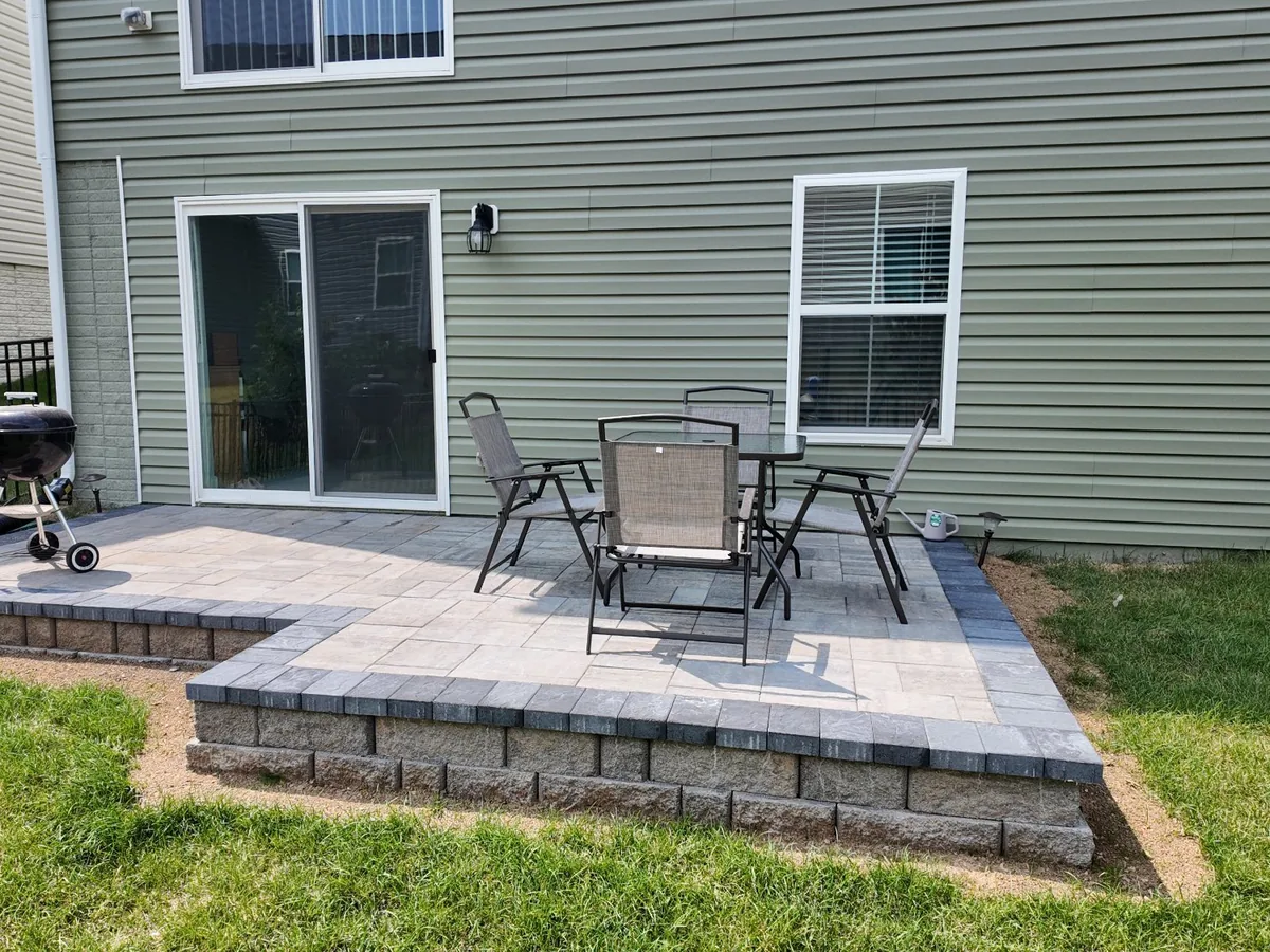 A raised gray paver patio on a block base behind a house with a grill and chairs, brick and stone patios