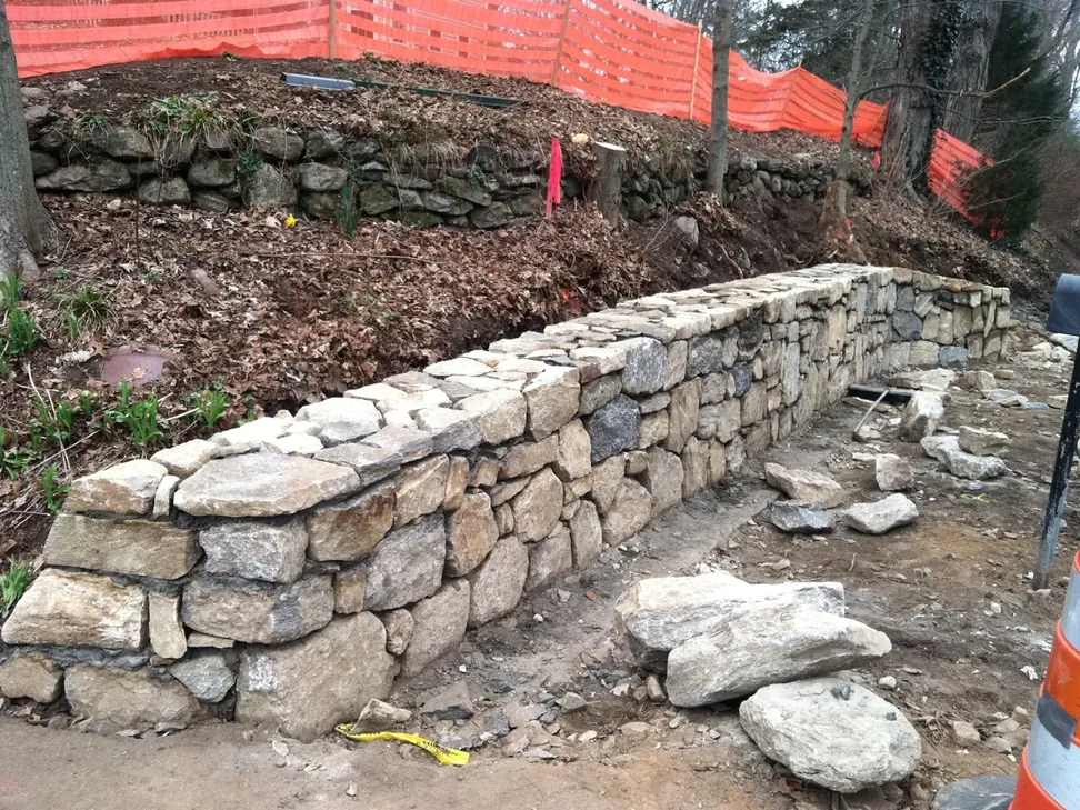 A dry laid natural stone retaining wall under construction along a wooded slope, retaining walls